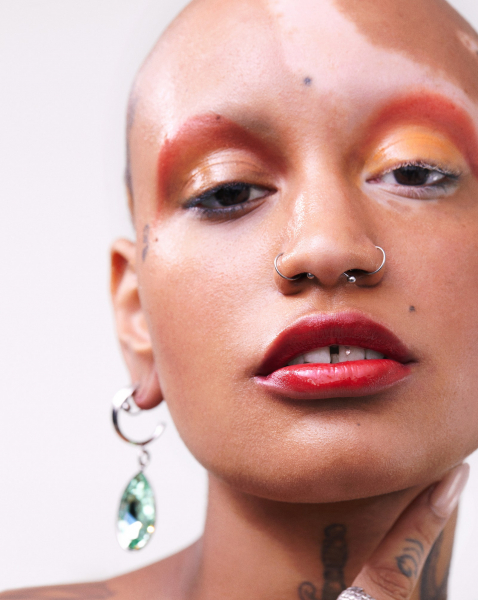 close up portrait of a bald person with two nose rings wearing orange eye shadow and red lipstick