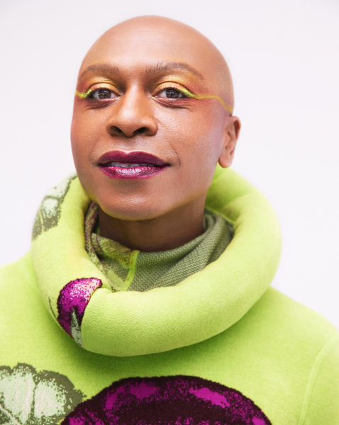 photo of a bald person slightly smiling into camera. they wear lime green winged liner and metallic purple lipstick that...