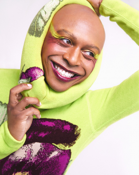 photo of a bald person smiling broadly while looking off camera. they wear lime green eye makeup and metallic purple...